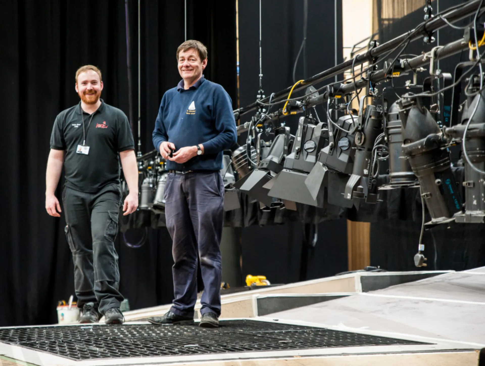 Two men in lanyards pose next to a lowered lighting rig