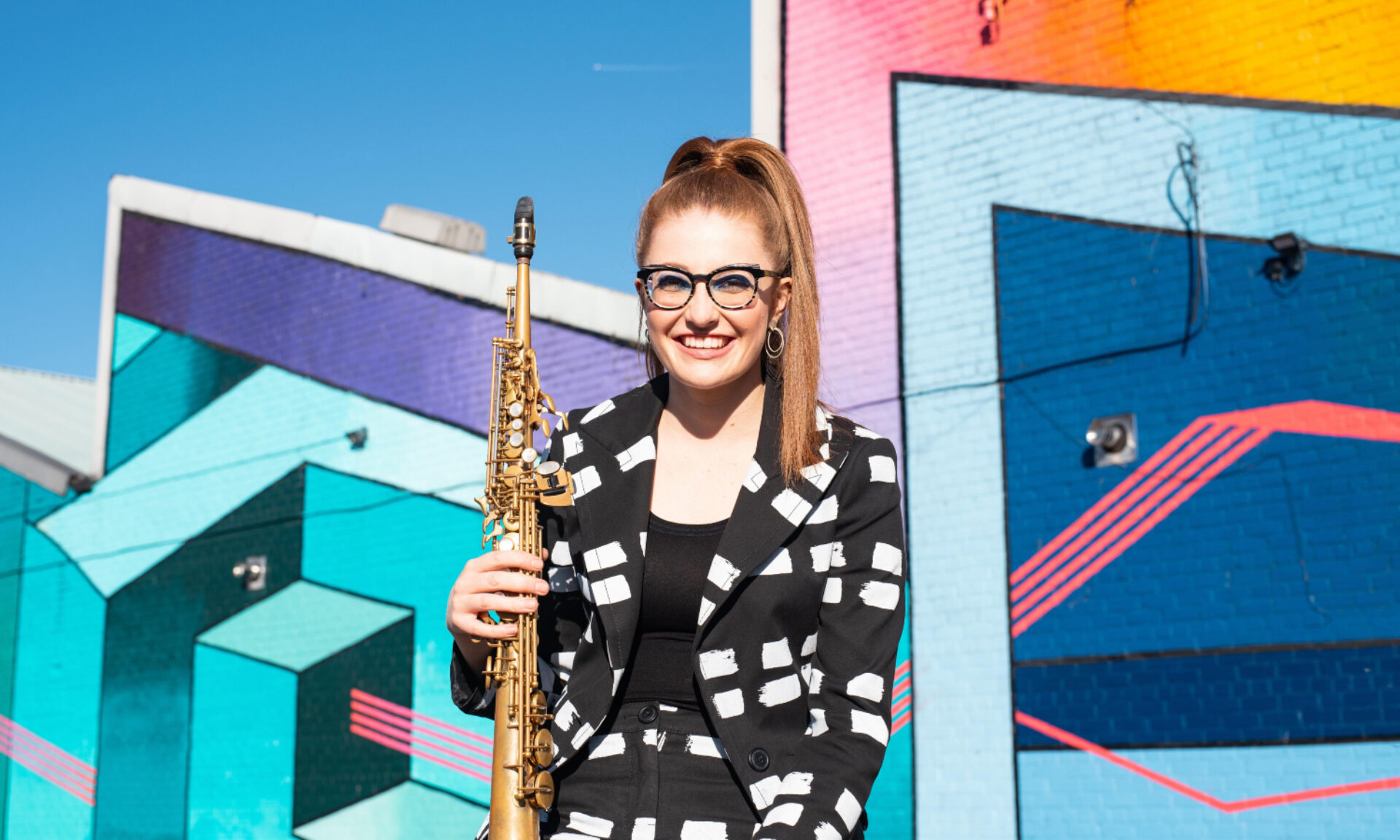 Jess Gillam holds her saxphone wearing a patterned monochrome suit in front of a bright graffiti'd wall